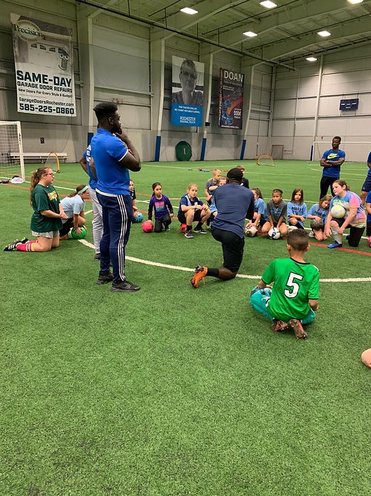 A large group of children seated on turf, listening to a coach's instructions during an indoor session.