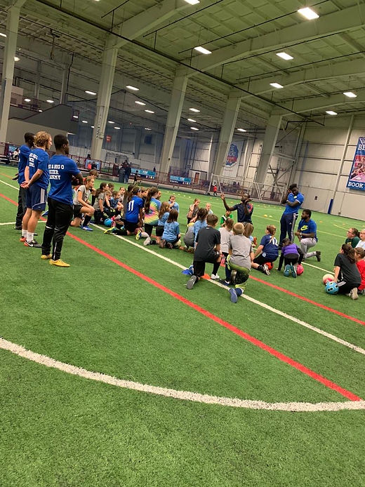 A young player dribbling a soccer ball across a wide indoor turf field while a coach observes.